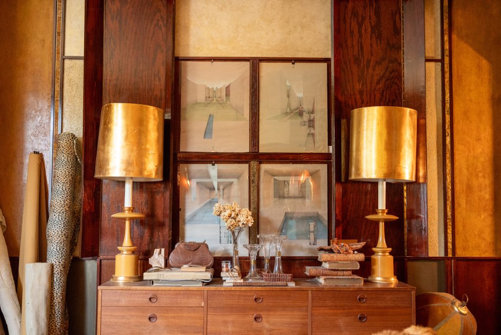 Image: A still life photograph of a room in the Greystone Home and Studio. At the center is a wooden credenza in front of a wood panel wall. Two golden lamps sit on each edge of the credenza. At the center of the credenza are old books, glass vases, a leather purse on top of large books, and dry flowers in a glass vase. On the left side of the credenza are rolls of fabric in various colors. Photograph by Tonal Simmons.