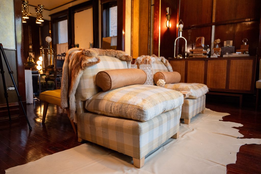 Image: An interior photograph of a room in the Greystone Home and Studio. Two light colored checkered armchairs with beige bolster pillows and furs wrapped around the back of the chairs sit at the center of the room on top of a cowhide rug. Photography by Tonal Simmons.