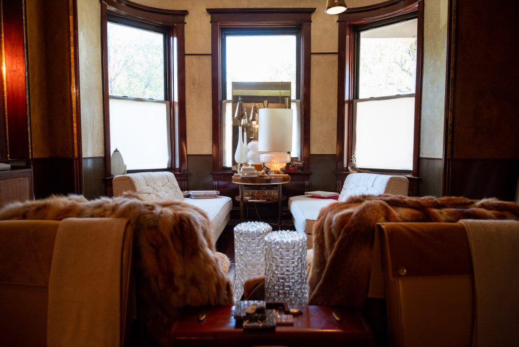 Image: An interior photograph of a room with three large windows. Two armchairs sit underneath two windows with a wood table in the center. Photography by Tonal Simmons.