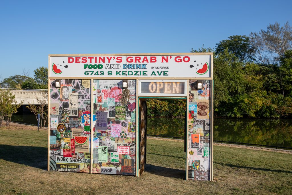 Image: Destiny’s Grab n’ Go installation by Carlos Flores and Deon Reed. The structure looks like a cornerstore facade. The top has a sign that says "DESTINY'S GRAB N' GO FOOD AND DRINK BY US FOR US 6743 S KEDZIE AVE". The rest of the facade is covered in layers and layers of posters, images, photos, and more. There is a sign above the doorway that says, "OPEN". Photo by Ruby Que. 