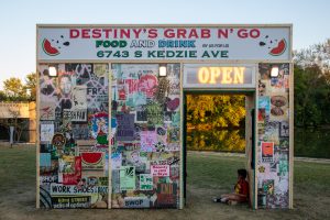 Imagen: Instalación Destiny 's Grab n' Go, de Carlos Flores y Deon Reed. La estructura parece la fachada de una tienda de barrio. En la parte superior hay un cartel que dice «DESTINY'S GRAB N' GO FOOD AND DRINK BY US FOR US 6743 S KEDZIE AVE». El resto de la fachada está cubierta de capas y capas de carteles, imágenes, fotos y más. Encima de la puerta hay un cartel que dice «ABIERTO». Foto de Ruby Que.