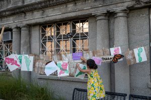 Image: Opening day celebration of diversión. A young child in a yello dress hangs up drawings on a string in Marquette Park. Photo by Ruby Que.