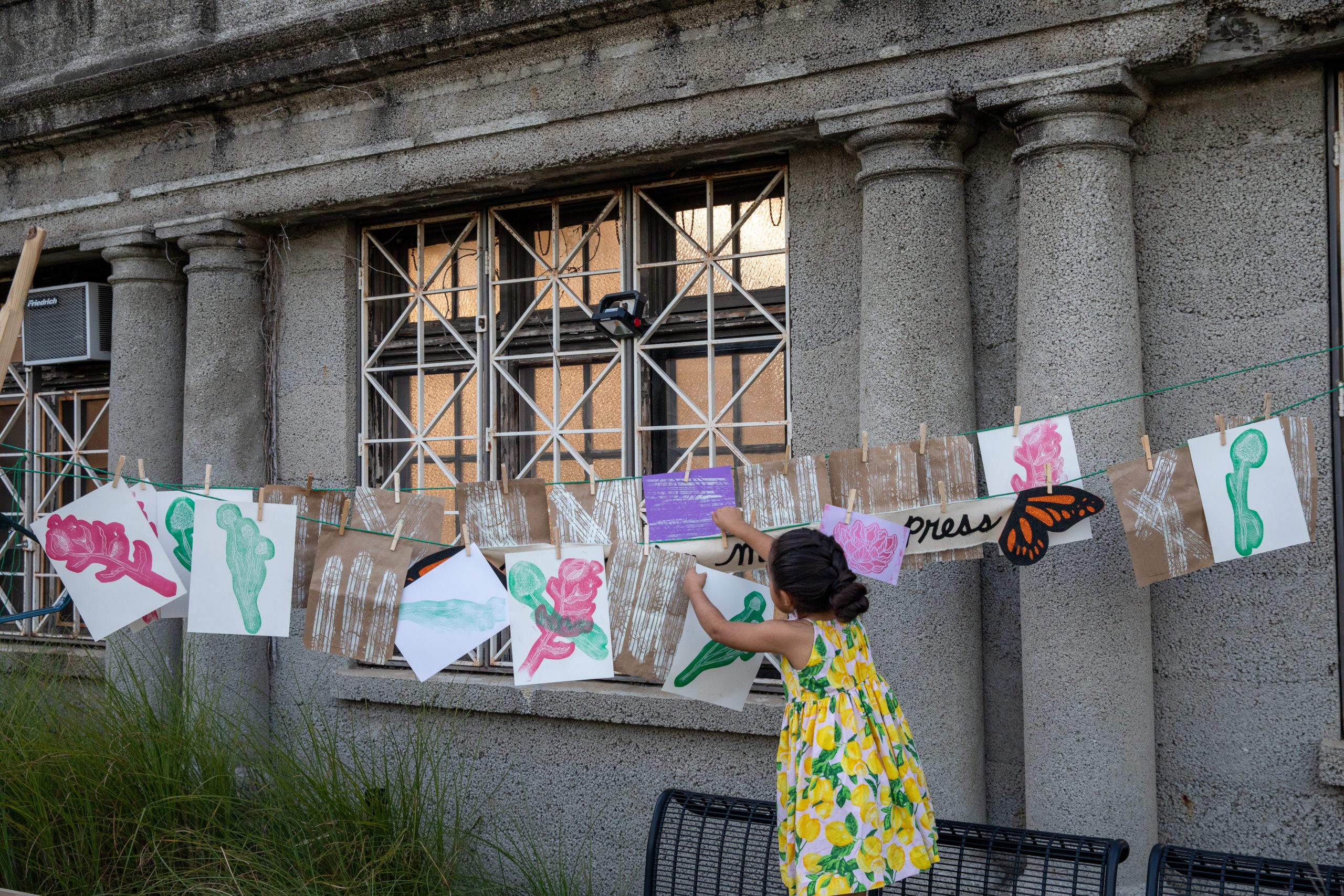 Image: Opening day celebration of diversión. A young child in a yello dress hangs up drawings on a string in Marquette Park. Photo by Ruby Que.