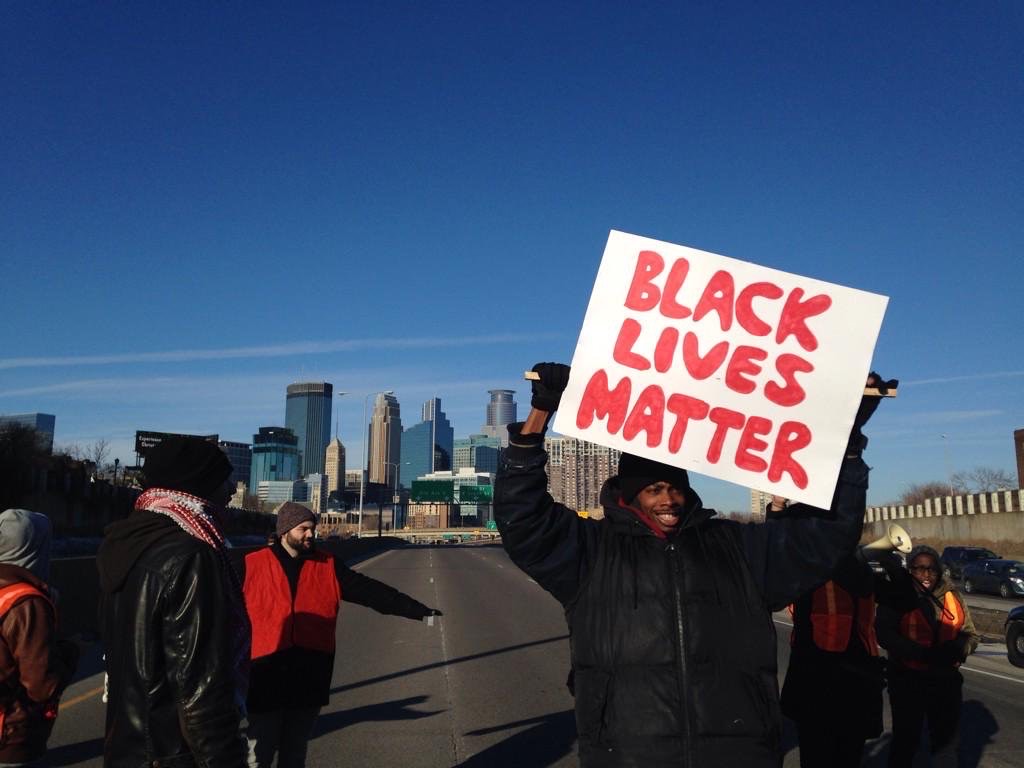 Image: A photo from a protest on Hwy 35w, circa 2015. A person in the foreground holds up a sign that says, “BLACK LIVES MATTER” in red. Photo by the author.