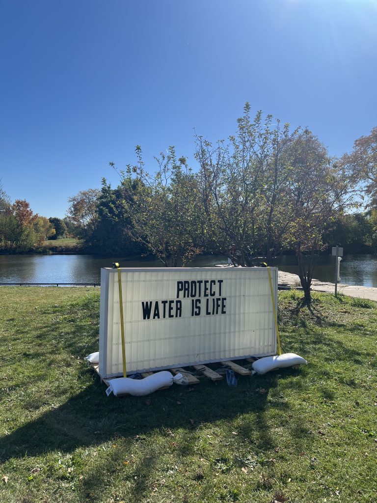 Imagen: Vista de la instalación de la obra Is This It, de Titus Wonsey. La pieza es un cartel de marquesina que dice «PROTEGER EL AGUA ES VIDA». Detrás del cartel se ve un lago. Foto de Cristobal Alday. 