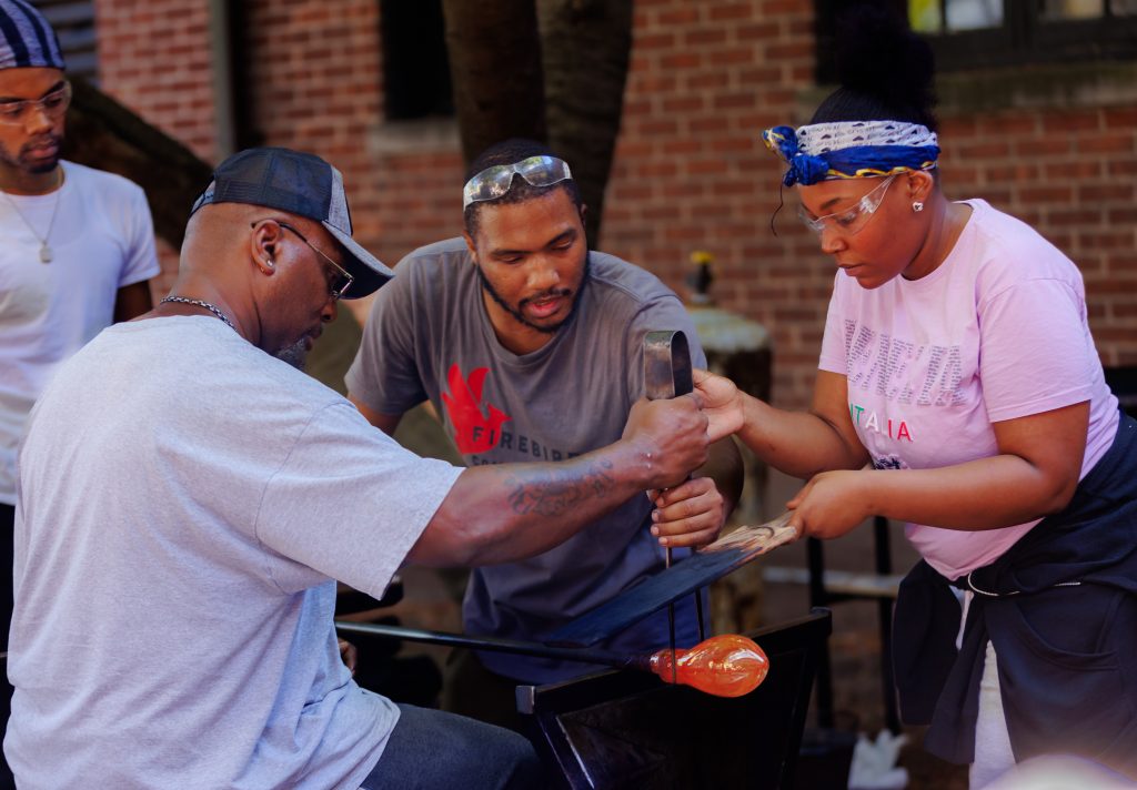 Image: This series of skills-sharing workshops between Firebird Community Arts and Red Line Service culminated in a glass blowing event. The educators of Firebird Community Arts taught the artists of Red Line Service to blow glass on the Hull-House courtyard, creating cups and paperweights. The Firebird instructor with his glasses on his forehead is N'Kosi and the glassblower in the baseball cap is Red Line Service artist James. Photographed by Sarah Larson.