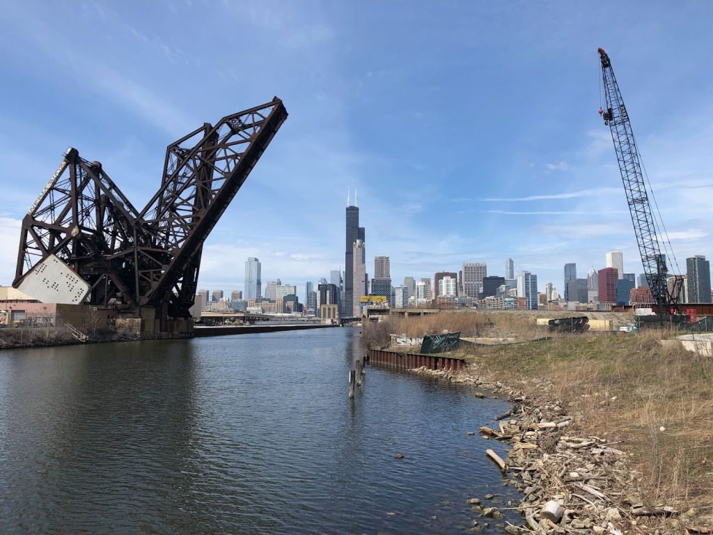 Image: Still image from Jane Buyck’s Chicago par ses riviéres / Chicago by its rivers, 2023, which shows a view  near the Chicago river's shore as the river is raised and the Chicago skyline looms behind the river on a clear day. 