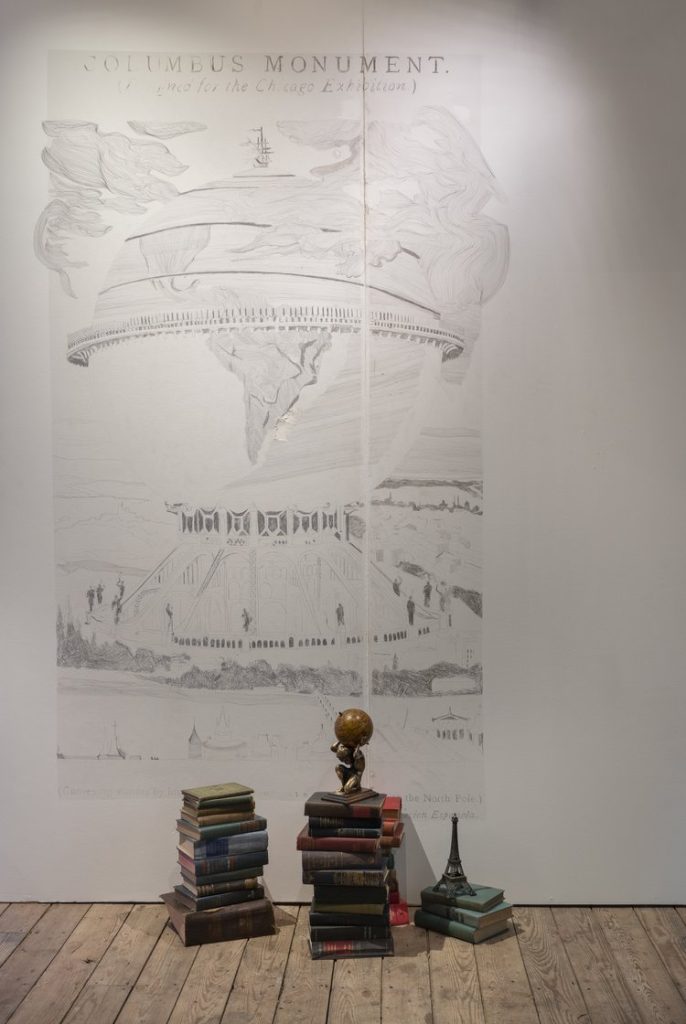 Image: Emilio Rojas, Memorial to an Unbuilt Monument and/or A Litany of Reduction (2017-2018). Durational drawing on wall from EXPO Chicago 2017 with 1892 World Columbian Exposition Memorial Silver Half Dollar. Three stacks of books of varying heights are placed in front of a pencil drawing of the Colombian exhibition on the wall behind them. A small figure of the Greek god Atlas is laid on top of the stack of books in the middle and a figure of the Eiffel Tower is laid on the small stack of books at the right. Photo courtesy of 6018|North.