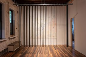 Image: Installation view of Aleksandra Walaszek’s “And If This is Home, Welcome Home,” 2024. Bronze-colored steel blinds shown open, light streams through the blinds, drawings can be seen on the walls around the installation. Image courtesy of 6018|North.