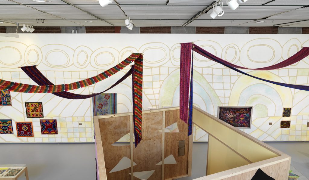 Image: Installation view of The United Colors of Robert Earl Paige, Hyde Park Art Center, 2024. A birds-eye view of an exhibition. A faint geometric mural covers the rear wall of the room. The wall is decorated with numerous artworks. Four long colorful textiles are hung from the ceiling, the tail ends dropping into an open wooden structure at the center of the room. Photo by Tom Van Eynde.