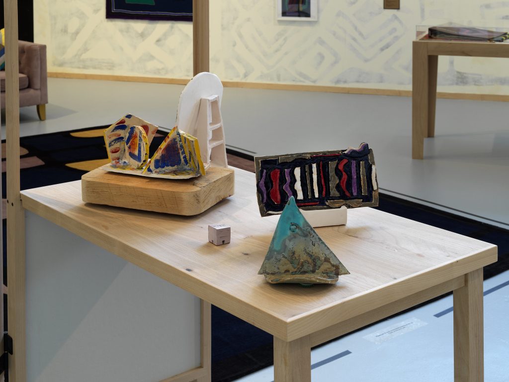 Image: Installation view of The United Colors of Robert Earl Paige, Hyde Park Art Center, 2024. A close-up image of several multicolored artworks displayed atop a wooden table. There is a geometric rug and a couch visible in the background. Photo by Tom Van Eynde.