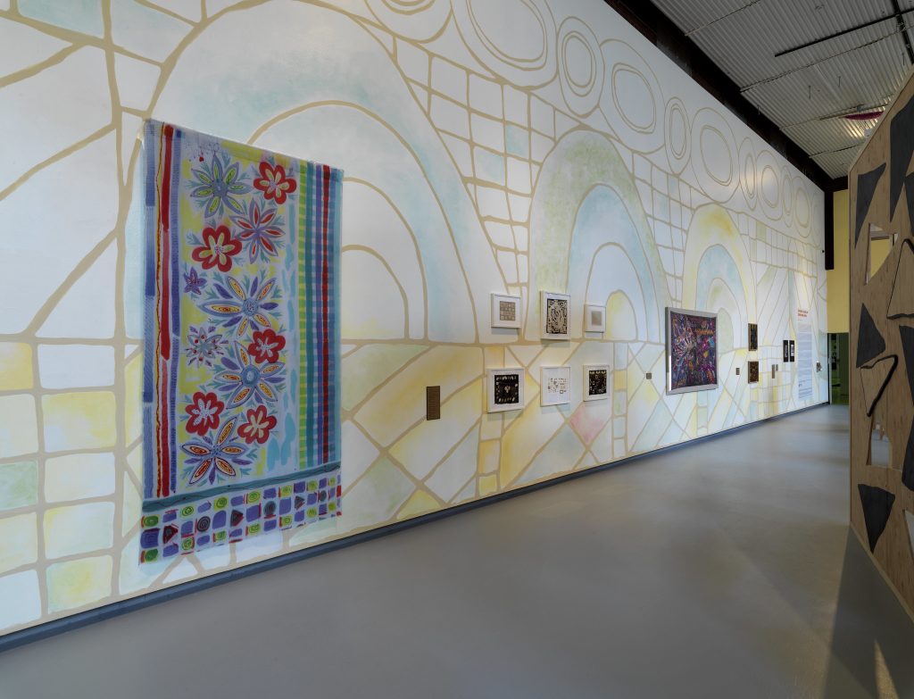 Image: Installation view of The United Colors of Robert Earl Paige, Hyde Park Art Center, 2024. A faint geometric mural covers the length of a wall. A multicolored textile decorated with floral designs is near the left side. There are numerous artworks displayed along the length of the wall. A structure with cutouts is slightly visible on the right side. Photo by Tom Van Eynde.