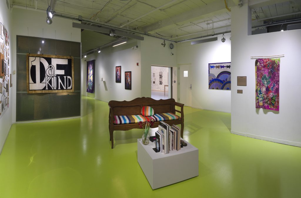 Image: Installation view of The United Colors of Robert Earl Paige, Hyde Park Art Center, 2024. A wooden couch with a multicolored cushion is displayed near the center of a small room. Artworks are hung along the walls of the hallway, including a hand-painted textile that reads "1 of a Kind." A display table in front of the couch is decorated with flowers and a selection of books. The floor is neon green. Photo by Tom Van Eynde.