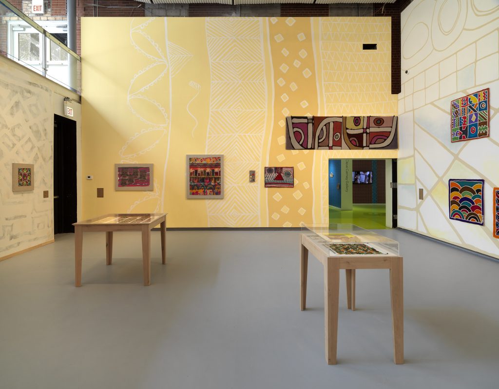 Image: Installation view of The United Colors of Robert Earl Paige, Hyde Park Art Center, 2024. Three walls are covered with faint geometric murals. The furthest mural is yellow. Each wall is decorated with numerous artworks. Two display cases are filled with artwork, one to the left and the other to the right side of the room. Photo by Tom Van Eynde.