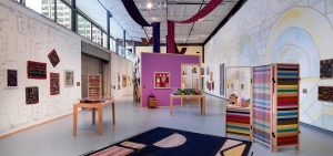 An installation view of an exhibition shows a geometric pattern rug in the foreground, a colorful room divider to the right, a large wall to the right with a faint, geometric mural with artwork displayed along the length of the wall. The opposite wall has a mural and artworks hung along it, intersected by a large garage door. Four long, colored textiles are draped from the ceiling, the tail ends dropping into a purple structure at the center of the room. There are two displays containing pillows and textile work.