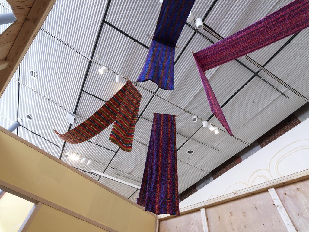 Image: Installation view of The United Colors of Robert Earl Paige, Hyde Park Art Center, 2024. Four multicolored textiles are hung from the ceiling, the tail ends dropping toward the viewer. The ceiling is metallic sheets intersected by lighting tracks. Wooden walls are visible near the bottom edge of the photo. Photo by Tom Van Eynde.