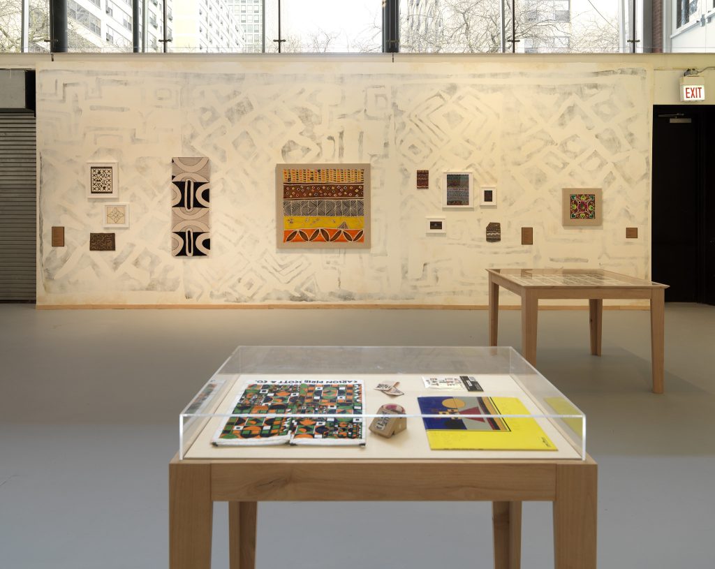 Image: Installation view of The United Colors of Robert Earl Paige, Hyde Park Art Center, 2024. A wall is covered with a faint geometric mural and decorated with numerous artworks. There is an open doorway on the right and a garage door on the left side of the image. There is a display near the open doorway and another in the foreground of the image. There are numerous artworks inside the display cases. Photo by Tom Van Eynde.