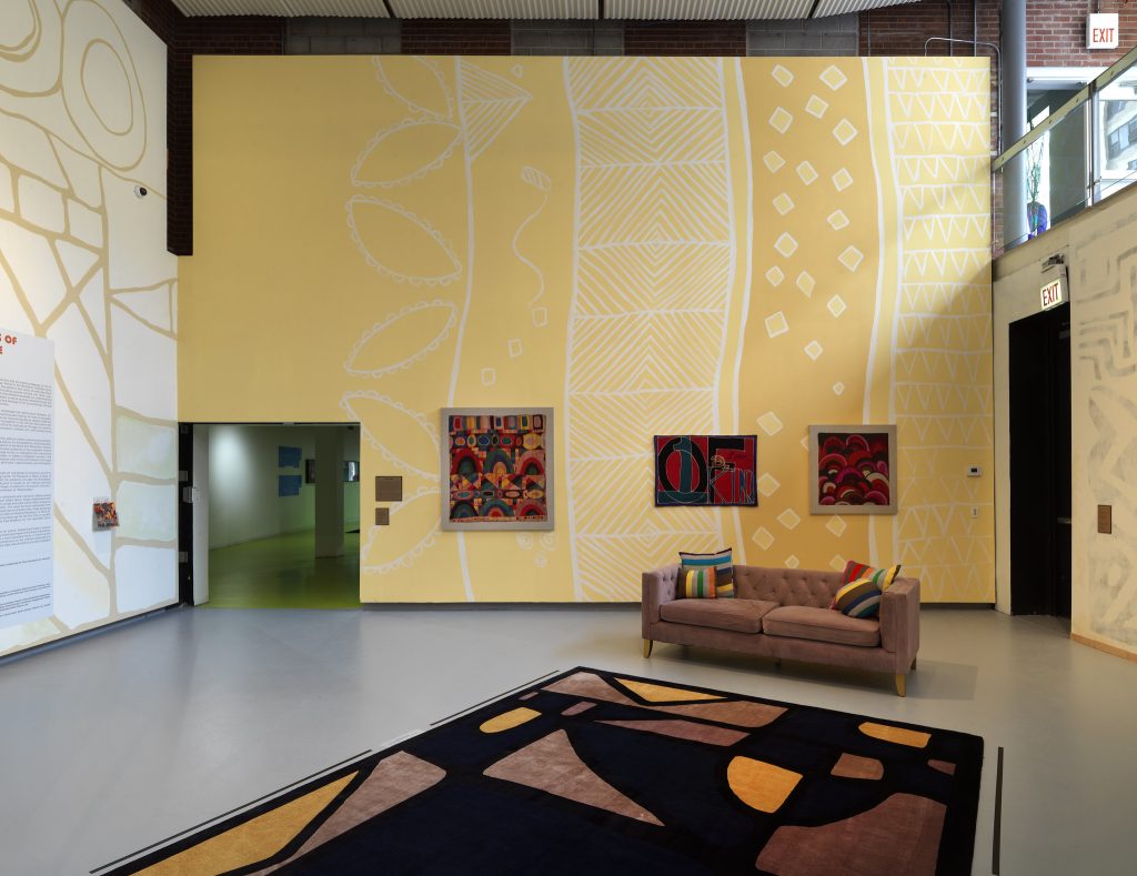 Image: Installation view of The United Colors of Robert Earl Paige, Hyde Park Art Center, 2024. A large rug with a yellow, black, and brown geometric pattern fills the foreground of the image. A brown couch with colorful pillows sits near the edge of the rug. The walls are covered with geometric murals. The wall furthest from the camera has vertical, yellow designs and is decorated with three multicolored artworks. Another exhibition room with neon green floor is visible through an open doorway on the left side. Photo by Tom Van Eynde.