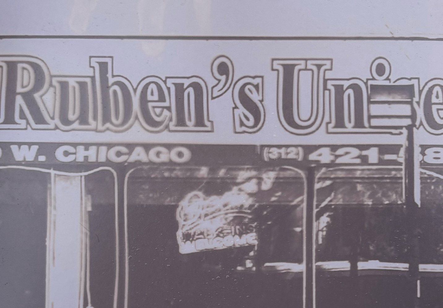 A window front with an awning that reads “Ruben’s” in a bold, outlined font, standing dark against the pale purple shade of the draping.