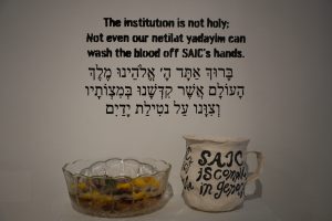 Image: Black text on a white wall reads, “The institution is not holy; Not even our netilat yadayim [Jewish ritual handwashing] can wash the blood off SAIC’s hands.” On a table in front of the text sits a glass bowl with water and flowers in it and a white jug with black writing stating, “SAIC is complicit in genocide.” Photo by Curators Under Censorship.