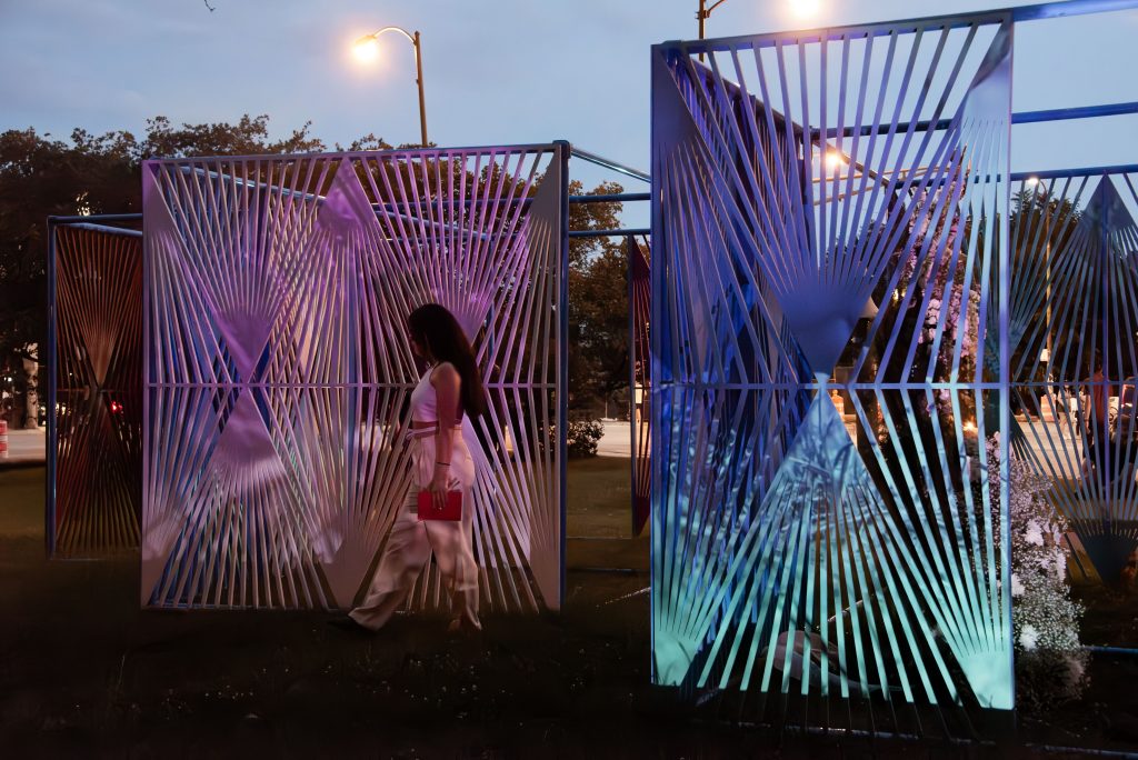 Image: Curator Ines Arango-Guingue walks between the multicolored latticework panels of Edra Soto’s La Casa de Todos (Everyone’s Home). She is wearing all white and carrying a notebook. Indistinct images are projected onto the white and blue latticework panels and a wedding gown made from flowers is visible behind one of the screens on the right side of the image. Image credit: Maria Burundarena.