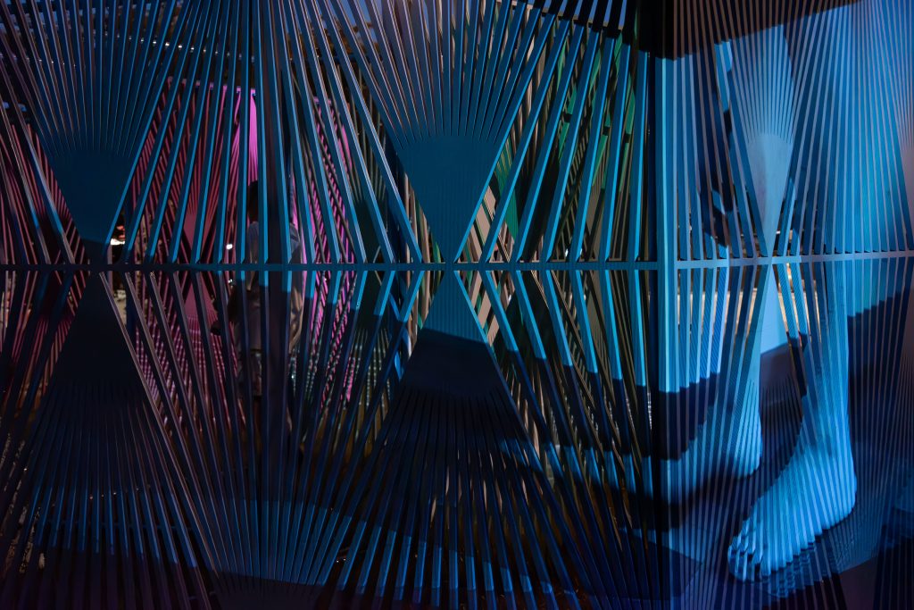 Image: An image of two feet from the knee down is projected onto Edra Soto’s La Casa de Todos (Everyone’s Home). The feet face toward the left side of the image, the foot furthest from the viewer distorted and lengthened as it wraps around the corner of the cerulean latticework screen. Pink, white, and green latticework screens are faintly visible through the abstract geometric pattern. Image credit: Maria Burundarena.