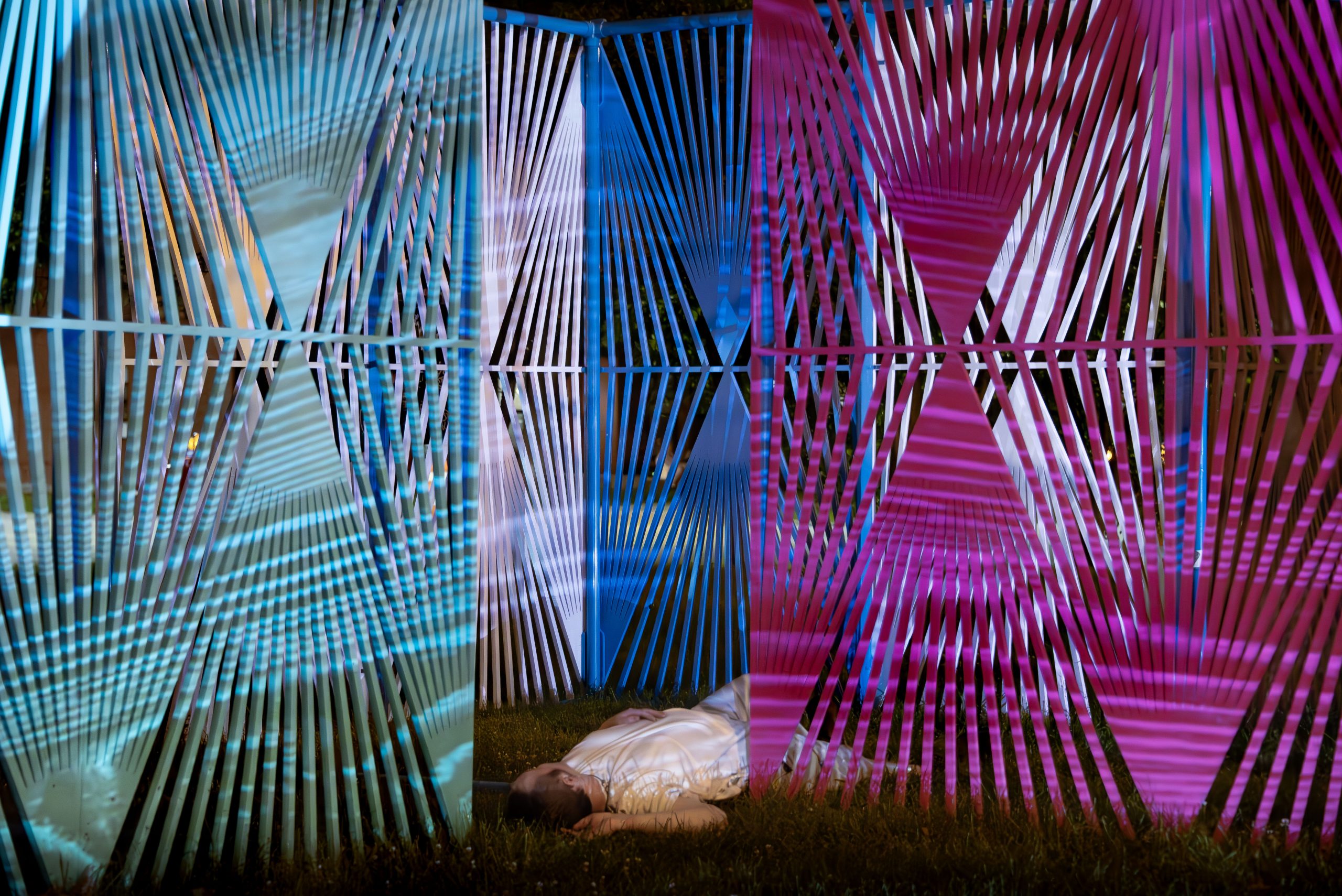 Performer Claire Fleming Staples lays supine on the grass between two colored, latticework panels with geometric patterns for "Suspira Es Hacia Adentro, Susurrar Es Hacia Afuera." Their legs are partially concealed by a pink panel on the right side. The panel to the right is cerulean. White and blue latticework panels are visible behind Staples.