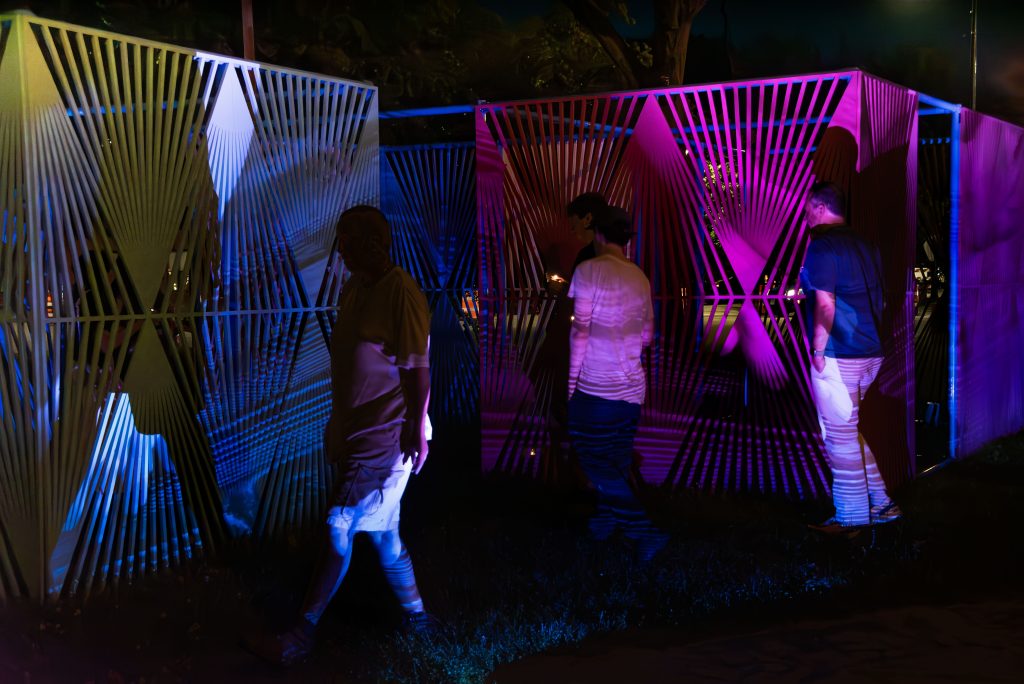 Image: Audience members walk through Edra Soto’s La Casa de Todos (Everyone’s Home) during Suspira Es Hacia Adentro, Susurrar Es Hacia Afuera. Three audience members stand in front of a pink latticework screen, indistinct images projected onto their figures and the screen behind them. A fourth audience member appears to be walking toward the left side of the image, casting a shadow of their figure on the white latticework screen. Image credit: Maria Burundarena.