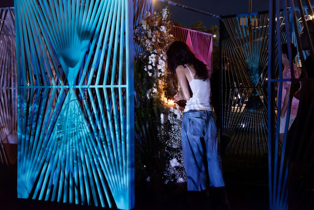 Image: An audience member lights a votive candle at the shrine constructed in the center of a wedding gown fashioned from white flowers. The gown is partially hidden behind a cerulean lattice work panel with an abstract geometric pattern. A projected image of a face is faintly visible on the cerulean panel. Image credit: Maria Burundarena.