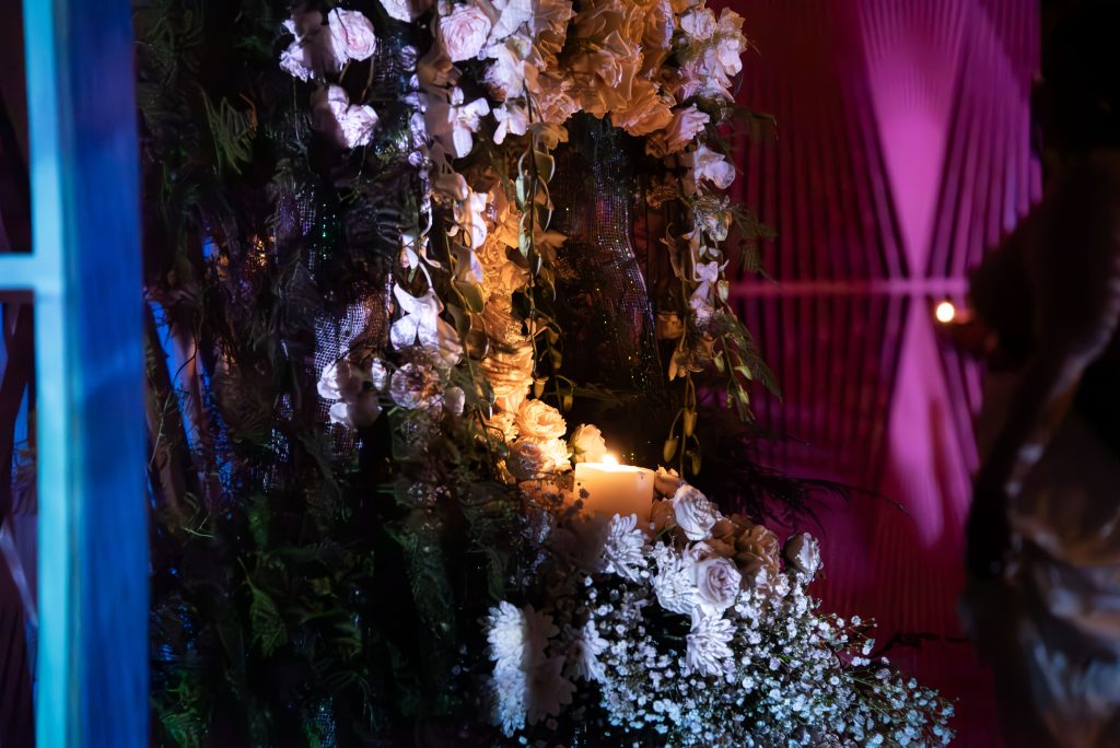 A detail shot of the wedding gown shrine constructed by Vince Phan. White baby’s breath and roses surround a burning candle. A pink latticework screen with an abstract geometric pattern is visible in the background.