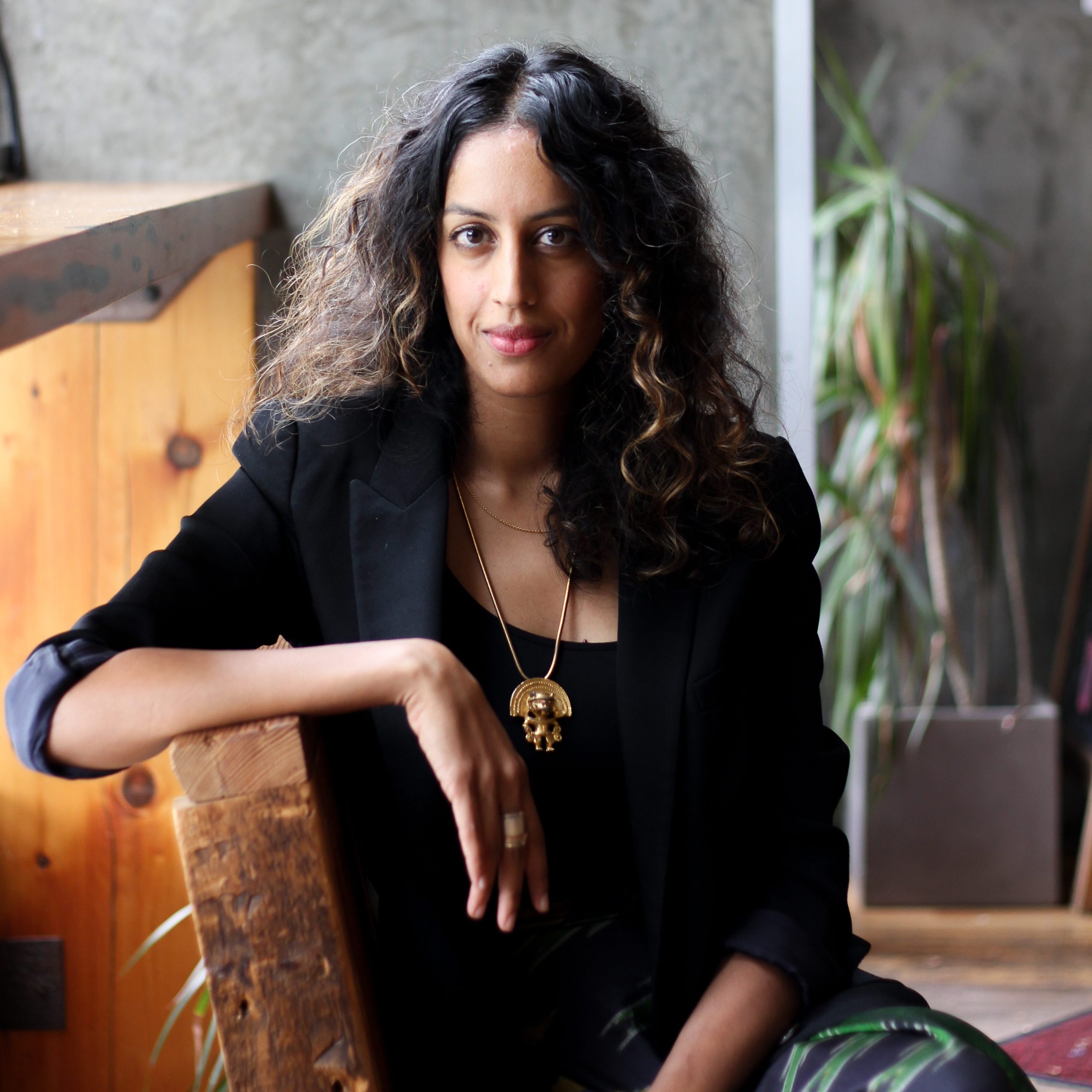 Ralapati has brown curly hair and is wearing a black blouse and green pants. She is pictured sitting sideways with her right arm resting on a wooden chair. She is seated in from of a tree light plant and  thick wood table.