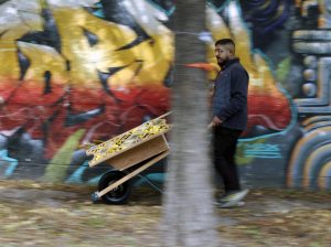 Image: Porta(til) Performance, video still by Ruby Que. Pushing a wheelbarrow in front of a graffiti wall. Courtesy of the curator.