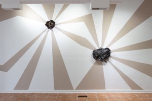 Image: A photograph of two brown ceramic pieces mounted, one higher than the other, on a white wall with brown clay stripes connecting them. To the left and higher up, The Protection and Collectivity of the Partridge Pea depicts a flower-like organism. Above With Those Dark Wings, lower to the right and slightly larger, is a bird-like creature.. The Protection and Collectivity of the Partridge Pea (2024) (left) 14x11x4 Wood Fired Ceramic, Missouri Clay Above With Those Dark Wings (In Awe) (2025) (right) 22x17x3 Wood Fired Ceramic, Missouri Clay. Courtesy of United Colors Gallery. 