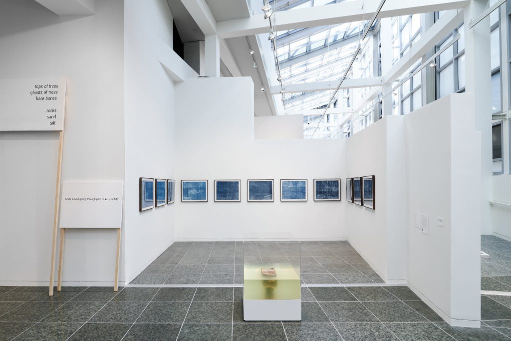 Image: installation view, the river runs slow and deep and all the bones of my ancestors / have risen to the surface to knock and click like the sounds of trees in the air, Wexner Center for the Arts, Columbus, Ohio, February 5th – July 9th, 2023, Photo Credit: Stephen Takacs. The piece Burn it, Bury it, Drown it can be seen in the foreground, created from a stack of Urdu primers bound and submerged in water. In the background are 11 mostly blue works on paper. They are all the same size.