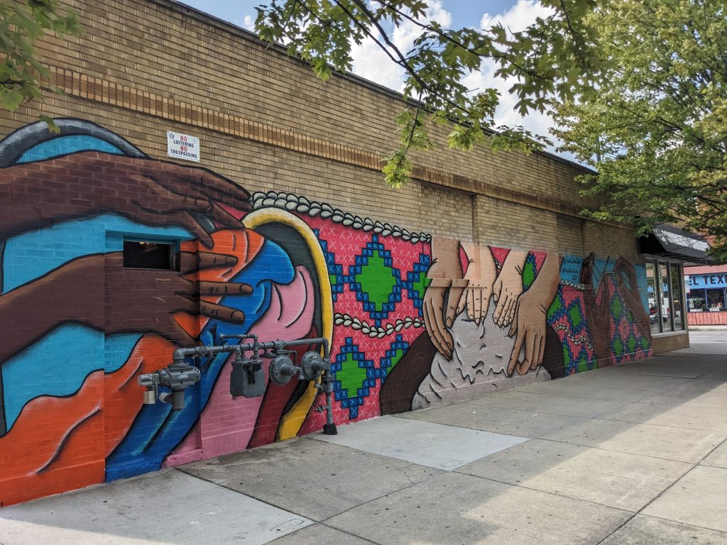 Image: Colorful Albany Park mural showing hands working on weaving, mending, sculpting, and other artistic activities. Image courtesy of Natalia Virafuentes.