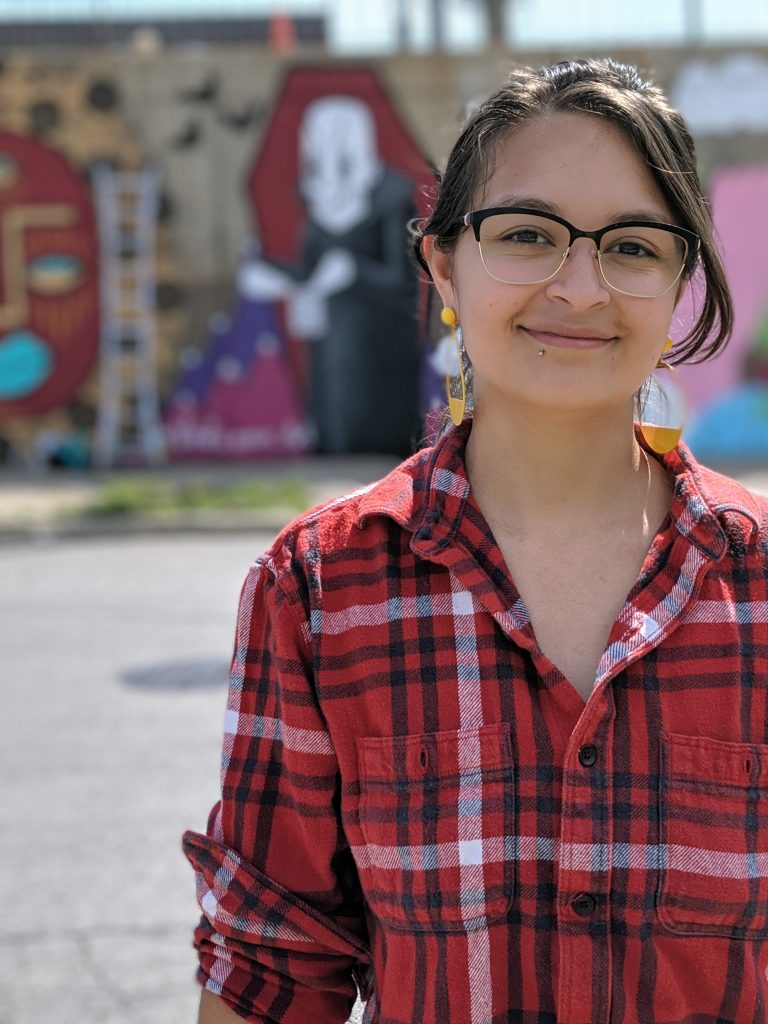 Image: Natalia Virafuentes stands in front of her wall mural wearing a red, white and black plaid shirt and yellow earrings. Photo courtesy of Natalia Virafuentes.