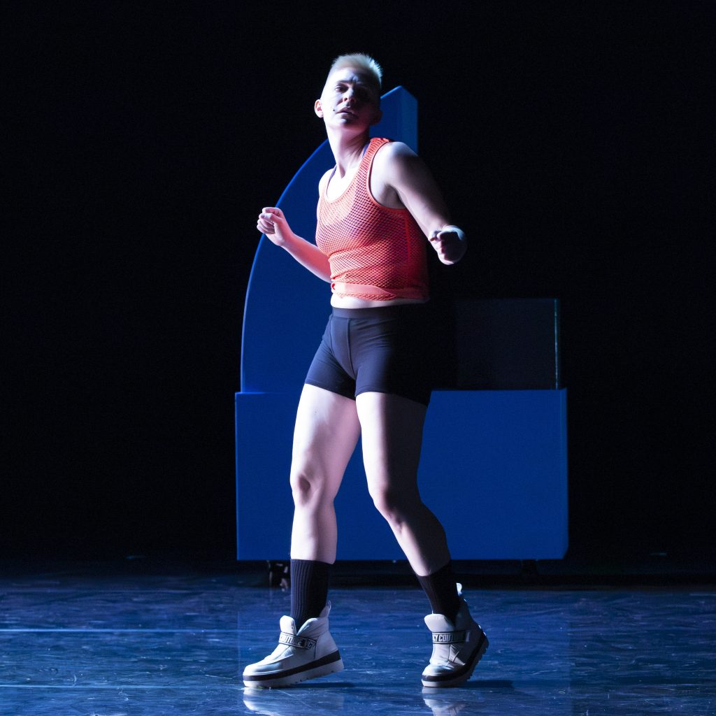 Image: Nora Sharp performing Cosmic Docks on March 12, 2025 at Chicago Solo Spotlight Festival, Dance Center of Columbia College Chicago. Sharp can be seen taking a boxer's stance alone on the stage under blue lighting.