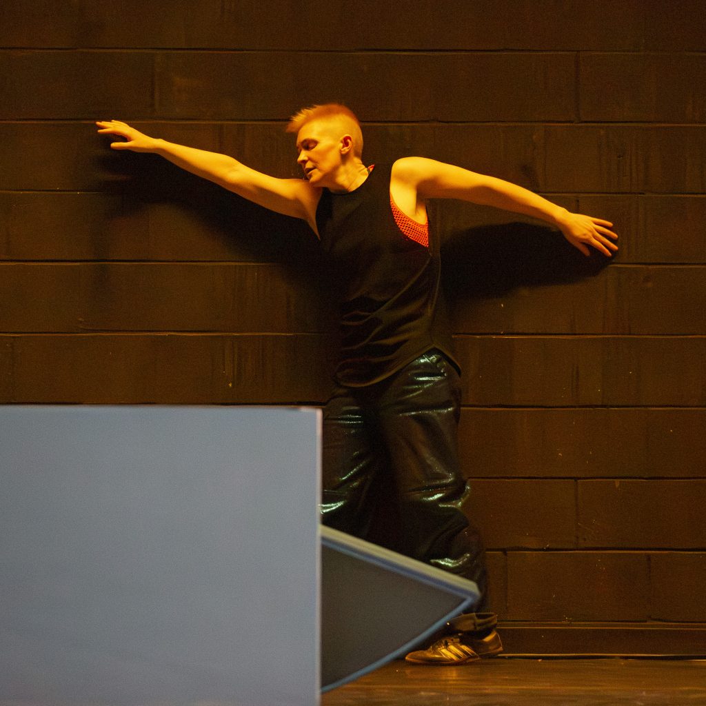 Image: Nora Sharp performing Cosmic Docks on March 12, 2025 at Chicago Solo Spotlight Festival, Dance Center of Columbia College Chicago. Sharp can be seen moving against the brick wall with a strained, exasperated look.