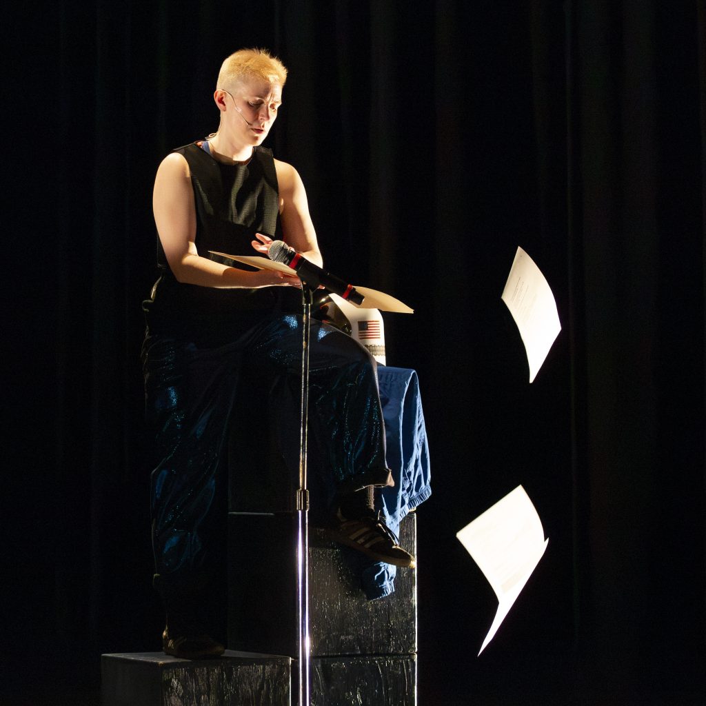 Image: Nora Sharp performing Cosmic Docks on March 12, 2025 at Chicago Solo Spotlight Festival, Dance Center of Columbia College Chicago. Sharp can be seen on a painted black stepping stool flinging pages of their script off the stage. Sharp's hands are near the microphone. 