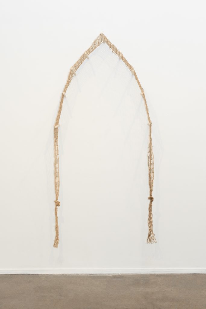 Image: Front view of ebere agwuncha, lancet portal featured in soft structures hung on a gallery wall at Chicago Artist Coalition, 2024. The 12 foot hand knotted fishing net with knotted ends drape over wood dowels that protrude from the wall, and from a lancet arch. Photo credit: Lyric Newbern.