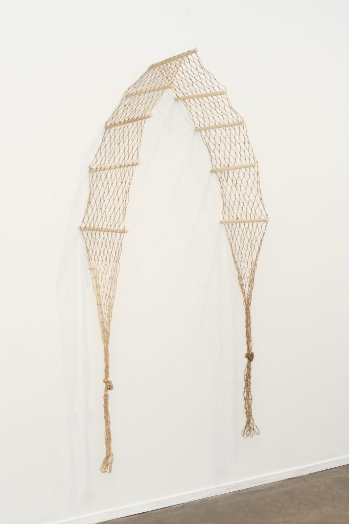 Image: Sideview of ebere agwuncha, lancet portal featured in soft structures hung on a gallery wall of Chicago Artist Coalition, 2024. The 12 foot hand knotted fishing net with knotted ends drape over wood dowels that protrude from the wall, and from a lancet arch. Photo credit: Lyric Newbern.
