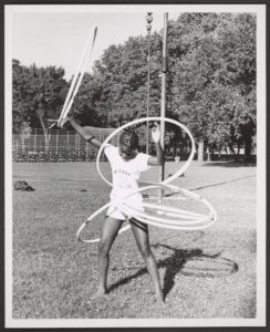 Image: A black and white photograph of a young African American girl skillfully playing with five hula hoops simultaneously on July 15, 1974. She stands in the grass at McKinley Park, with mature trees, the gate of a baseball field, and a tall pole in her background. She wears a short-sleeved shirt and shorts. Her hair is styled with pattern braids. Photo Credit: unknown, Courtesy of Chicago Park District Records: Photographs at the Chicago Public Library. Special Collections & Preservation Division, Digital Link.