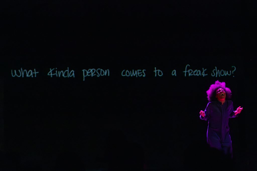 Image: Freeman | Po'Chop is shown yelling out to the audience under purple lighting. The question “What kind of person comes to a freak show?”, is hung behind her across the stage in blue hand-written text. Photo by William Frederking.