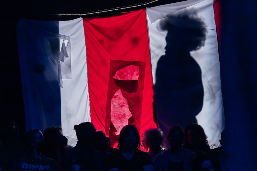 Image: View of the audience in front of the stage which shows a torn red and white vertically-striped flag. Freeman | Po'Chop's shadow is seen behind the flag. Image by Natasha Moustache.