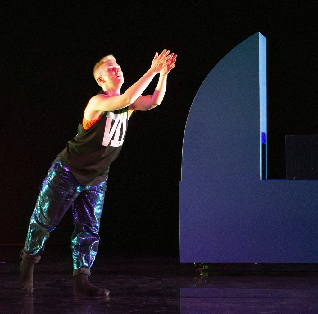 Image: Nora Sharp performing Cosmic Docks on March 12, 2025 at Chicago Solo Spotlight Festival, Dance Center of Columbia College Chicago. Sharp can be seen on a gesturing with her hands pointed above her. A piece piece of the stage can be seen behind Sharp.