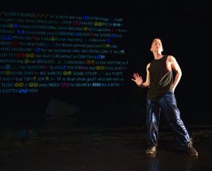 Image: A still from Nora Sharp's performance of "Cosmic Docks" on March 12, 2025 at Chicago Solo Spotlight Festival, Dance Center of Columbia College Chicago. Sharp can be seen wearing a black tank top and blue reflective pants while posing confidently in front of a paragraph consisting of a flirty copypasta text message on a black background. Image by William Frederking.