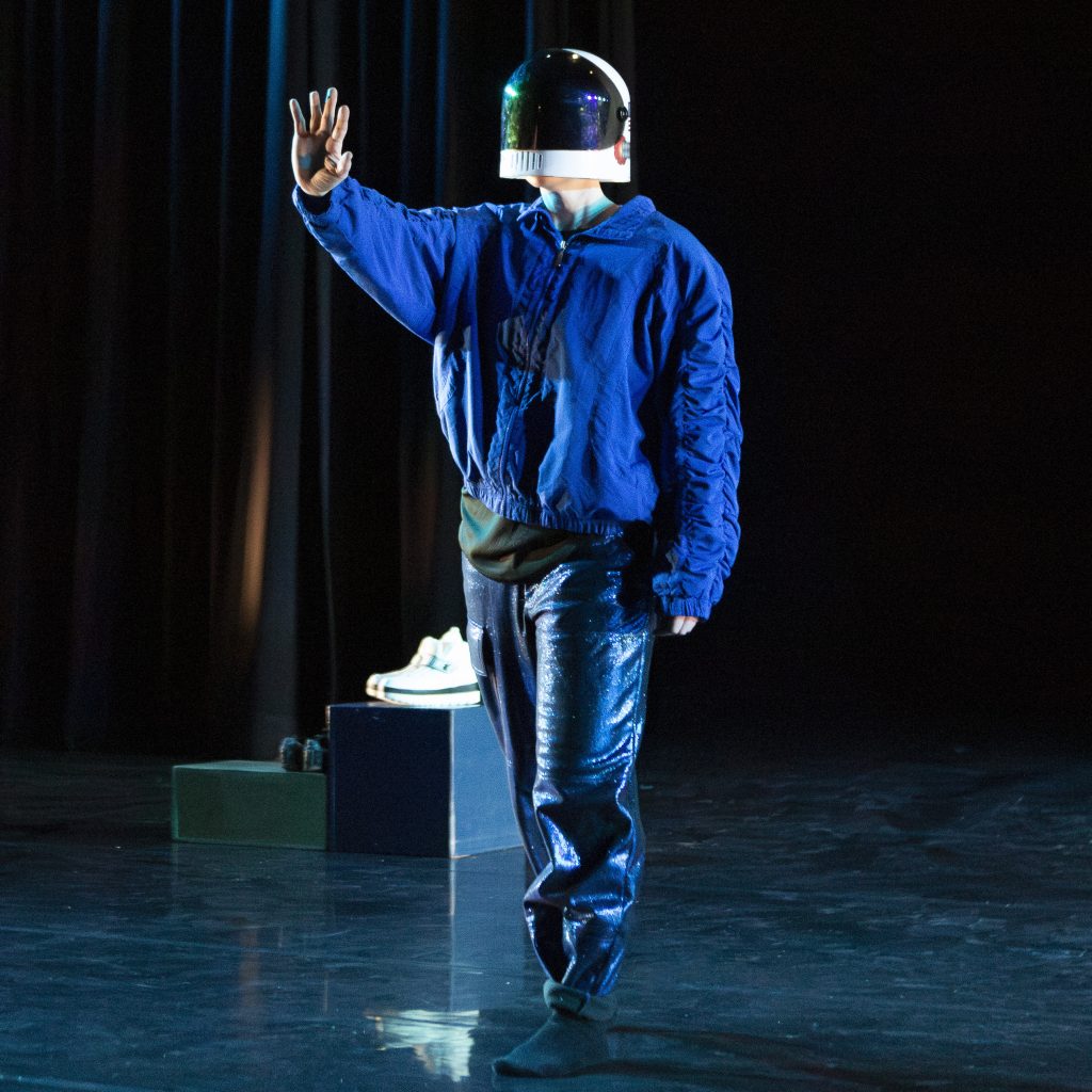 Image: Nora Sharp performing Cosmic Docks on March 12, 2025 at Chicago Solo Spotlight Festival, Dance Center of Columbia College Chicago. Sharp can be seen taking a step forward on the stage while wearing a motorcycle helmet. On both of the steps behind Sharp are shoes: the first step has black shoes and the second step has white sneakers. Sharp is wearing no shoes, just black socks.