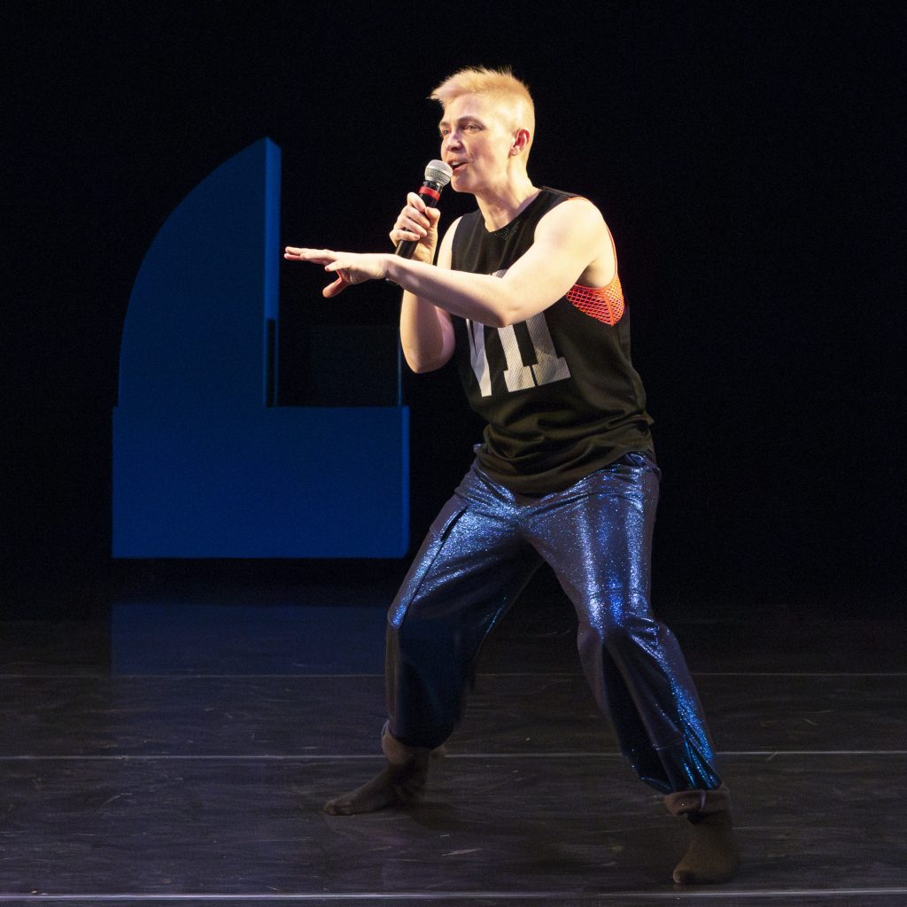 Image: Nora Sharp performing Cosmic Docks on March 12, 2025 at Chicago Solo Spotlight Festival, Dance Center of Columbia College Chicago. Sharp can be seen taking the stance of a stand-up speaking directly to the audience.