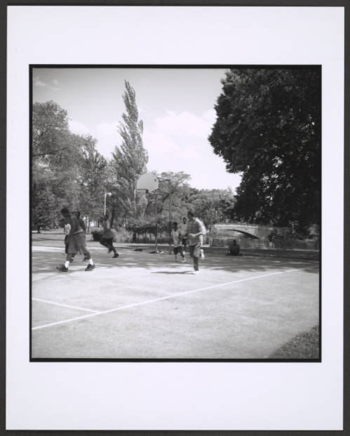Image: A black and white photograph of eight young Black boys on a basketball court. Park. To the right, one kid sits on the ground, while the other seven are captured in movement, seemingly in the midst of a basketball game. The drop is filled with mature trees, a walking bridge, and water. Photo Credit: Unknown date and photographer, Courtesy of Chicago Park District Records: Photographs at the Chicago Public Library. Special Collections & Preservation Division, Digital Link.