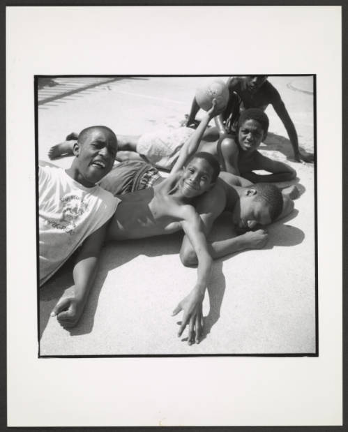 Image: A black and white photograph taken of five young Black boys lounging together on a paved surface, possibly by a pool in Sherman Park. The young boy in the middle lies on the paved ground, with his arms crossed and eyes closed, while the rest of the boys lean on and towards him. The boy to the left, positioned at the forefront, wears a white tank top, while a shirtless boy raises his arm playfully while holding a ball. Photo Credit: unknown date and photographer, Courtesy of Chicago Park District Records: Photographs at the Chicago Public Library. Special Collections & Preservation Division, Digital Link.